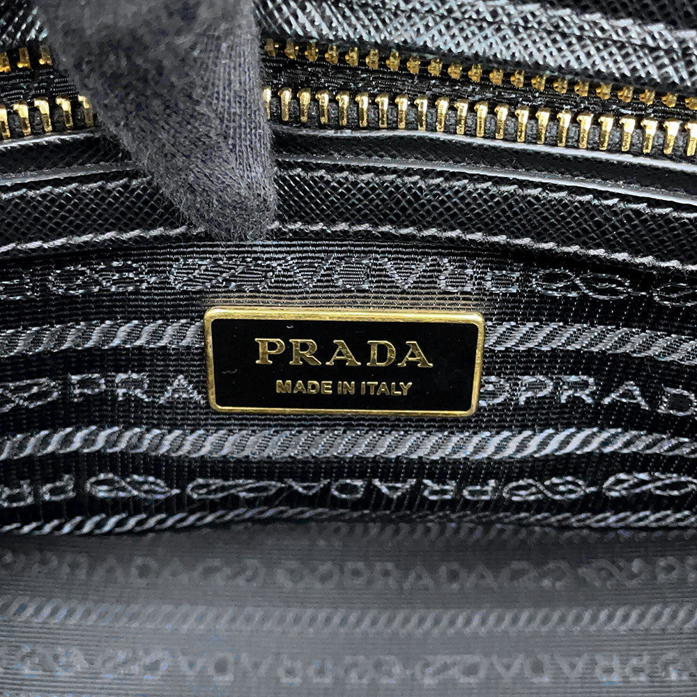 Close-up of a Prada interior label reading 'PRADA MADE IN ITALY' in gold on a black plaque, with a black and gray woven textile and gold zipper visible around it.