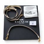 Coach Tabby shoulder bag shoulder strap laid out on its black branded box, with its white dust bag and care card also visible. The strap is light beige with dark trim and gold-tone hardware.