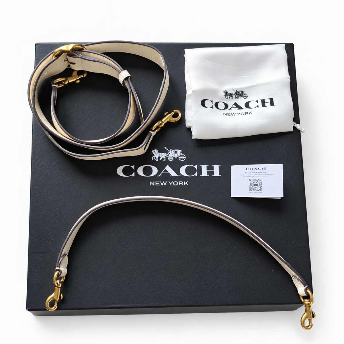 Coach Tabby shoulder bag shoulder strap laid out on its black branded box, with its white dust bag and care card also visible. The strap is light beige with dark trim and gold-tone hardware.
