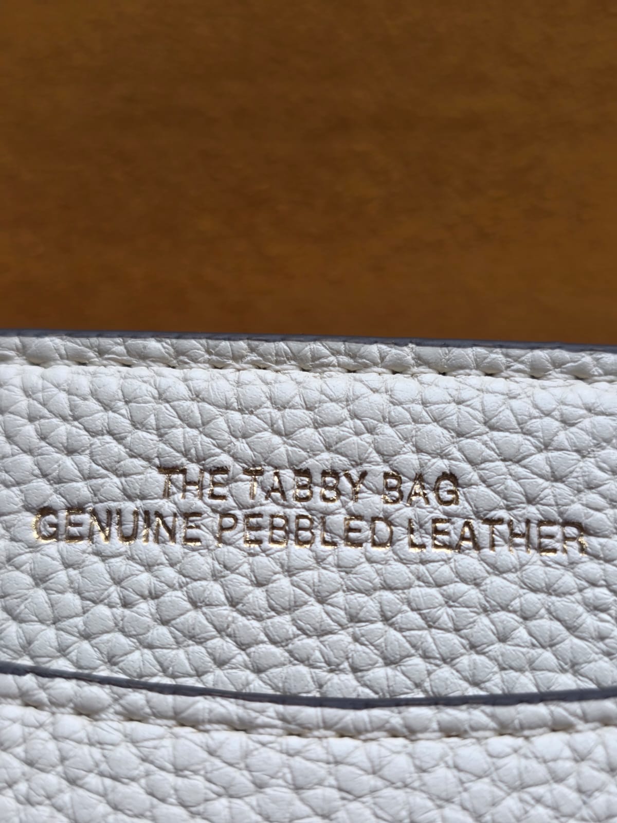 Close-up of a Coach Tabby 26 pebbled leather interior label with embossed text reading 'THE TABBY  BAG' and 'GENUINE PEBBLED LEATHER'