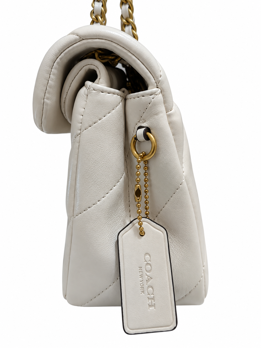 A close-up of a Coach quilted leather shoulder bag in off-white, showing the side profile with a gold-tone chain strap, a hanging leather tag, and gold hardware on a white background.